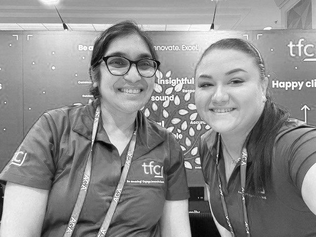 Harsha & Kira sitting together at a conference table, wearing TFC-branded collared shirts.