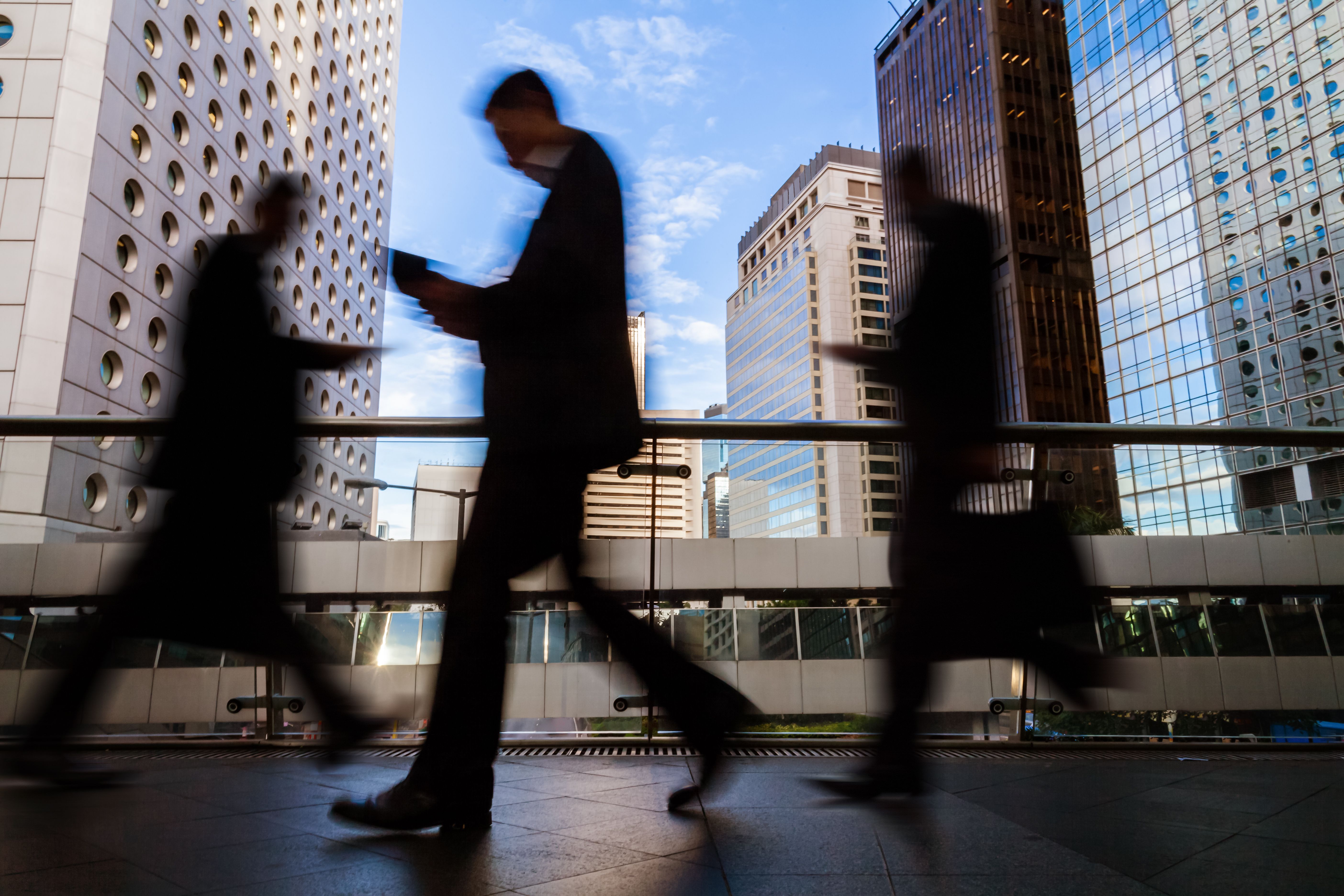 Silhouetted people in business attire walk past the camera, slightly blurred, while a downtown cityscape is visible in the background.