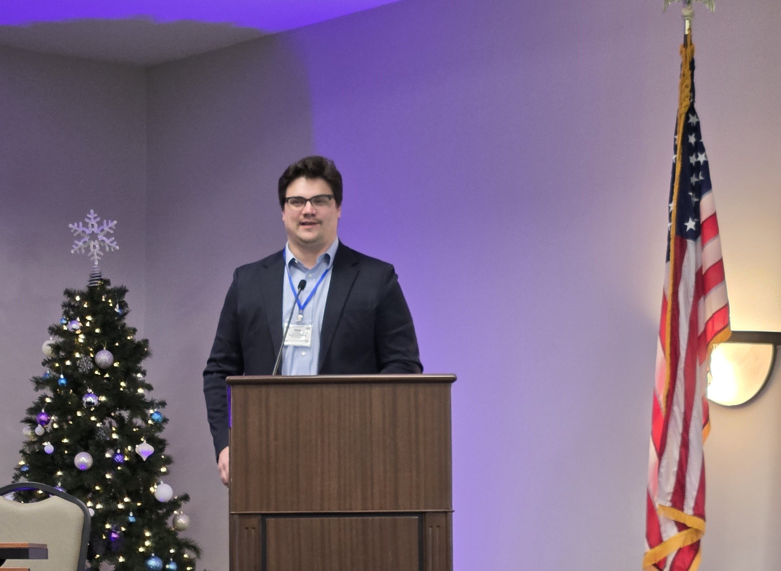 Nick gives an address at the AGA Commonwealth of Virginia Professional Development Training; he stands behind a wooden podium wearing a dark blazer and glasses. A Christmas tree, topped with a star, and an American flag are visible to either side of the podium.