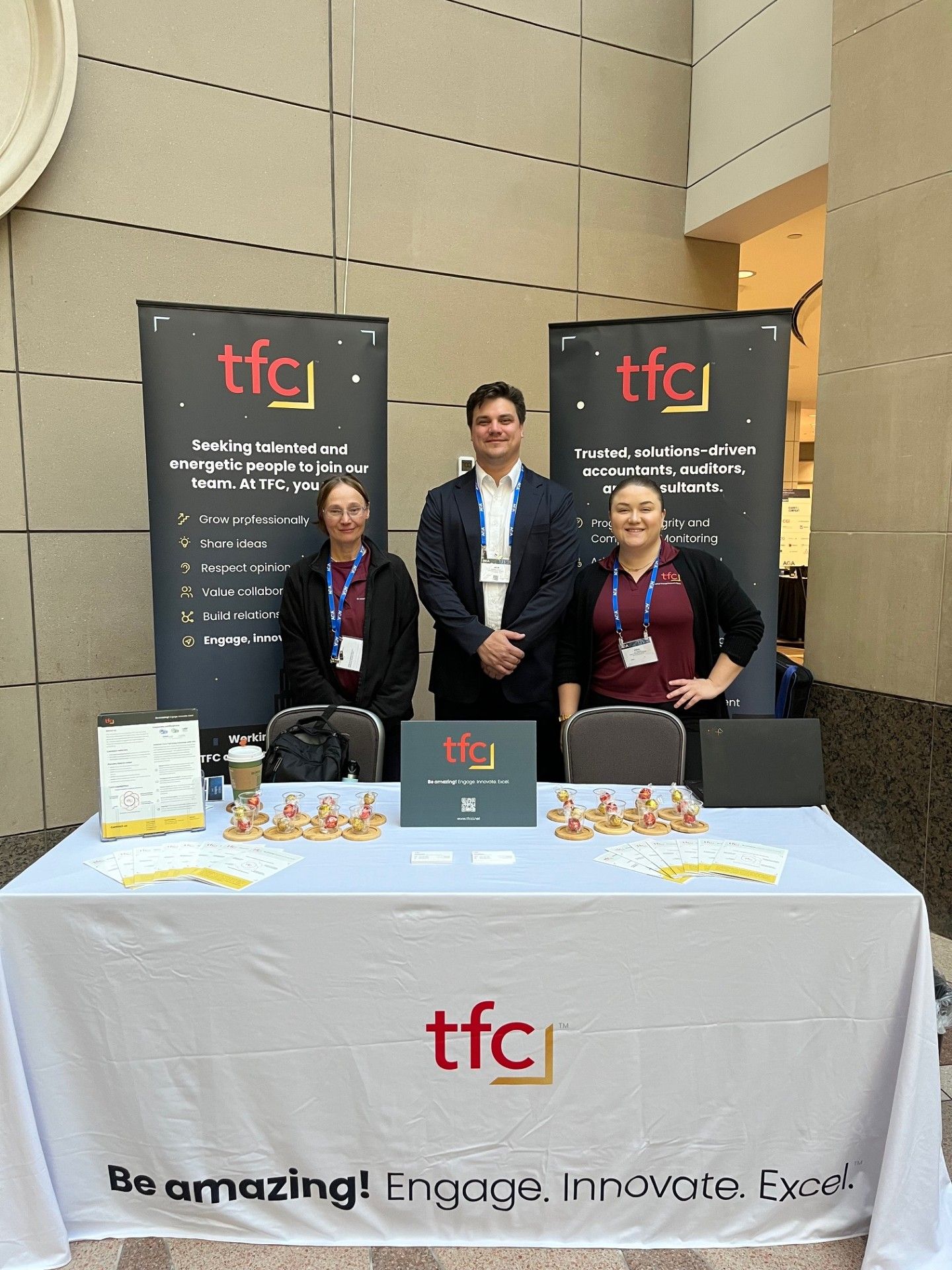 TFC team members stand together behind their table at the conference. Two banners with the TFC logo stand behind the table, while the table displays brochures, signs, and business cards.